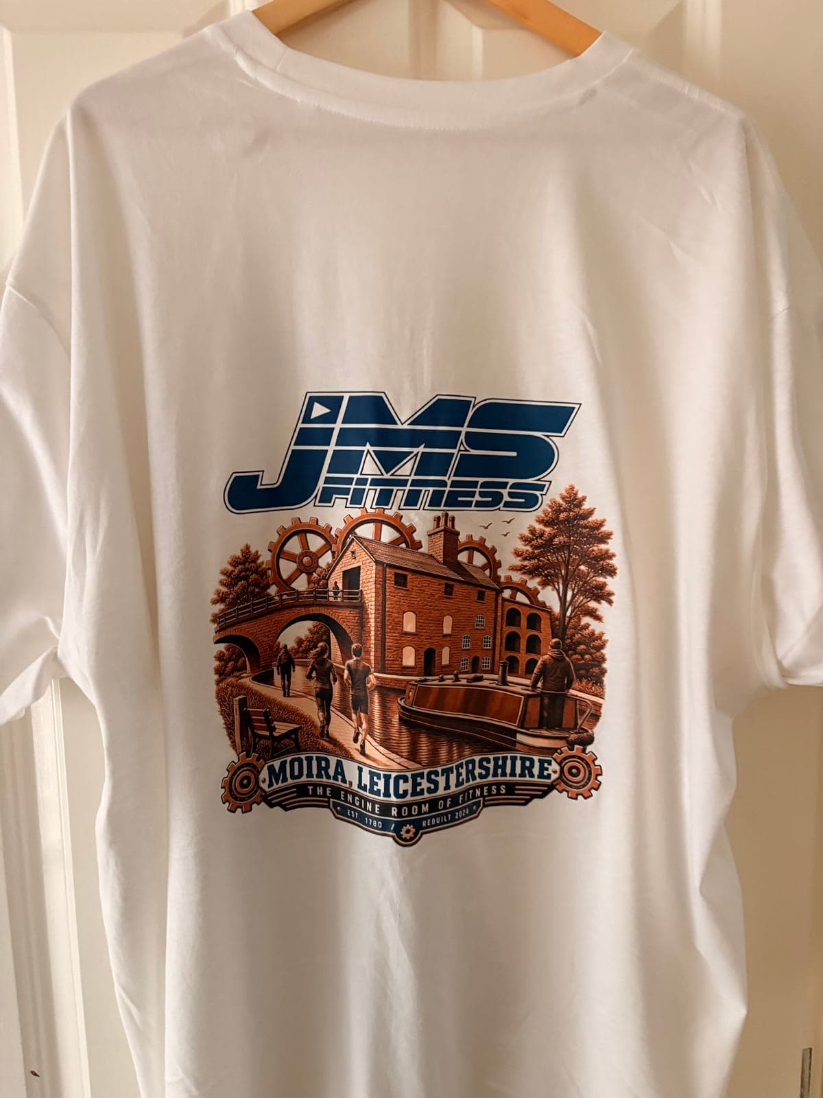 The back of the t-shirt: the JMS Fitness logo above an illustration of Moira Furnace with runners on the towpath and a narrowboat, over a banner reading Moira, Leicestershire — the engine room of fitness.